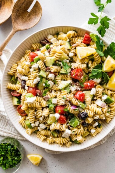 Greek pasta salad in a large white bowl with wooden serving spoons to the side.