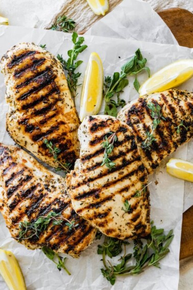 Four grilled chicken breasts on parchment paper with fresh herbs and lemon slices.