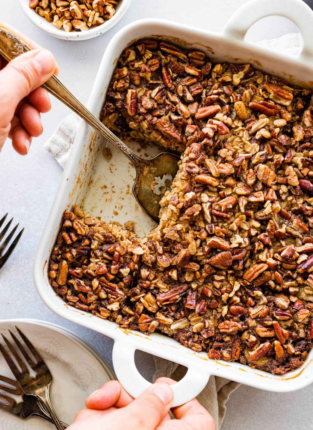 Baked oatmeal topped with chopped nuts in a white square baking dish with a woman's hand removing one slice.