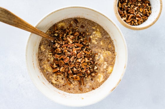 Chopped pecans added to the baked oatmeal batter in a white mixing bowl with a wooden spoon.