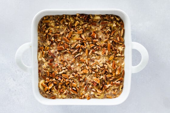 Unbaked baked oatmeal mixture topped with chopped pecans in a square white baking dish.