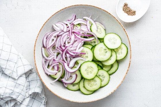 Sliced cucumbers and sliced red onion in a large white bowl.