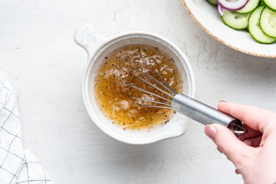 A woman's hand whisking a vinaigrette in a small white bowl with a metal whisk.