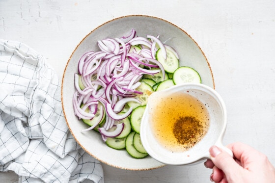 A woman's hand pouring a vinaigrette in a bowl of sliced cucumbers and sliced red onion.