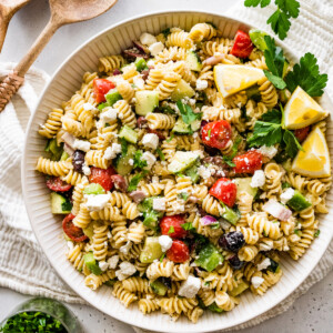 Greek pasta salad in a large white bowl with wooden serving spoons to the side.