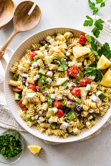 Greek pasta salad in a large white bowl with wooden serving spoons to the side.