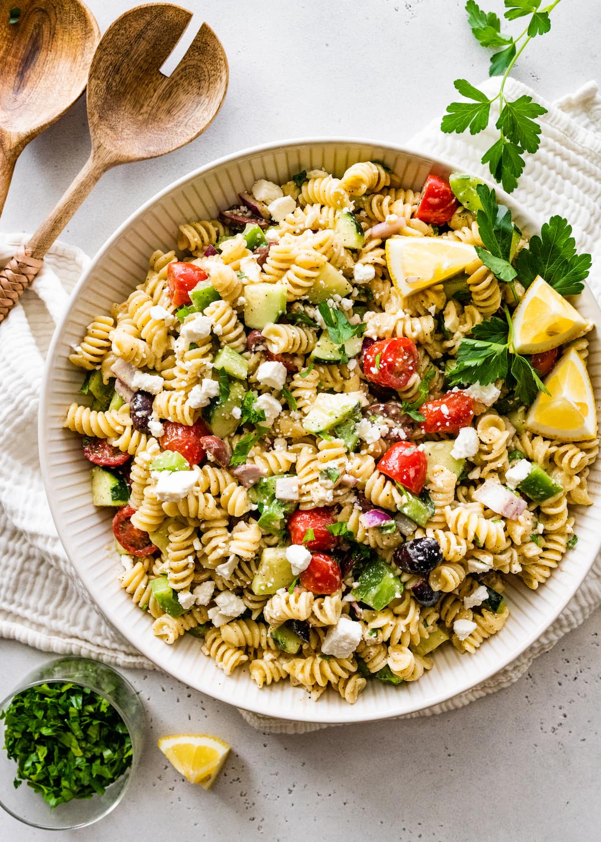 Greek pasta salad in a large white bowl with wooden serving spoons to the side.