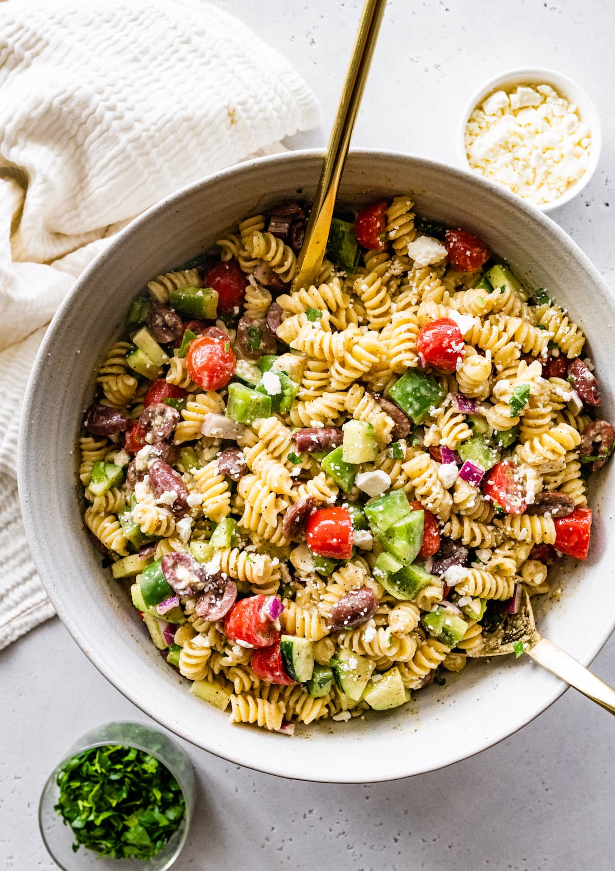 Greek pasta salad in a large white bowl with two metal serving spoons.