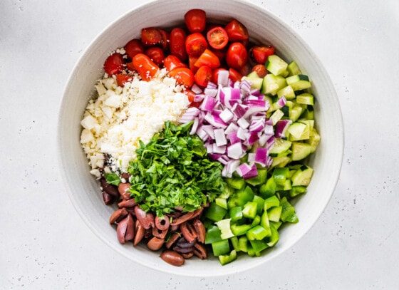 Greek pasta salad in a large white bowl. Noticeable ingredients include olives, green bell pepper, red onion, cucumber, cherry tomatoes, feta cheese, and parsley.