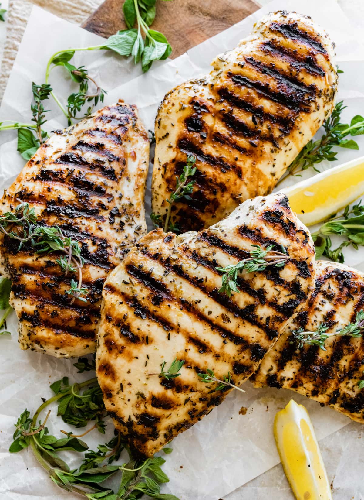 Four grilled chicken breasts on parchment paper with fresh herbs and lemon slices.
