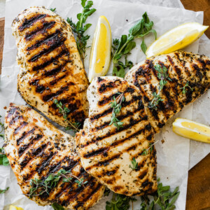 Four grilled chicken breasts on parchment paper with fresh herbs and lemon slices.