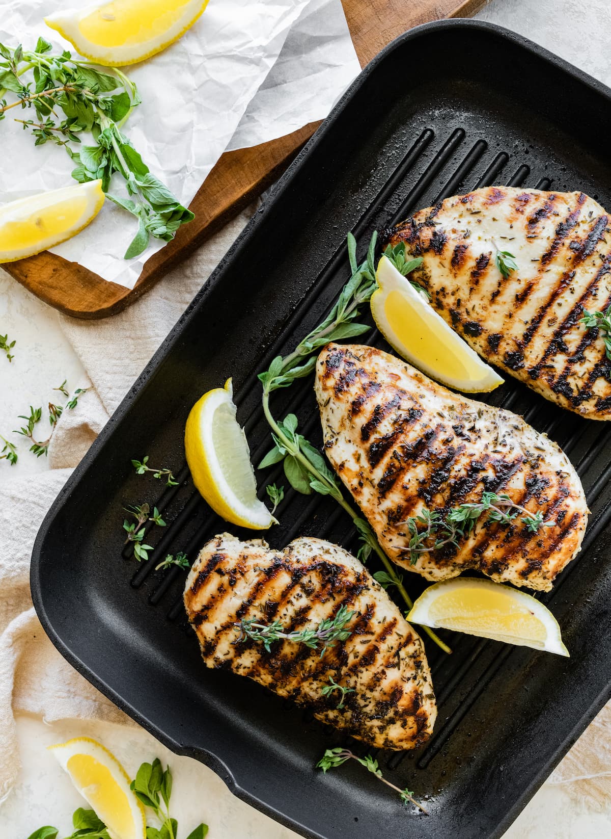 Three grilled chicken breasts in a grill pan with fresh herbs and lemon slices.