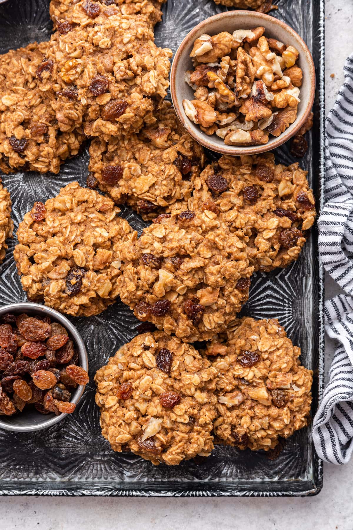 Multiple oatmeal breakfast cookies on a baking tray with two small bowls; one with walnuts and the other with raisins.