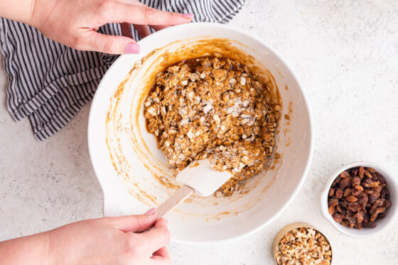 A woman's hand using a silicone spatula to mix the dough for the oatmeal breakfast cookies in a large mixing bowl.