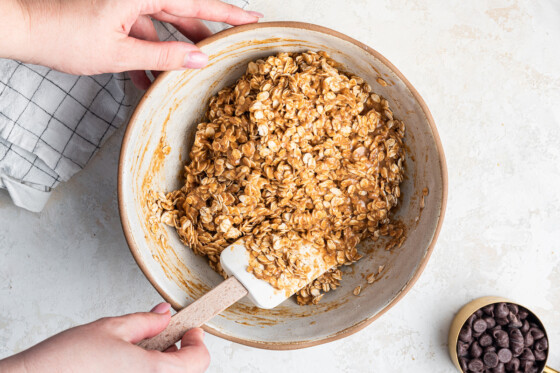 A woman's hand with a silicon spatula mixing the ingredients for oatmeal chocolate chip bars in a large mixing bowl.