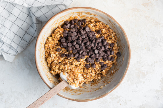 Ingredients for the oatmeal chocolate chip bars in a large mixing bowl with a silicone spatula and a cluster of chocolate chips.