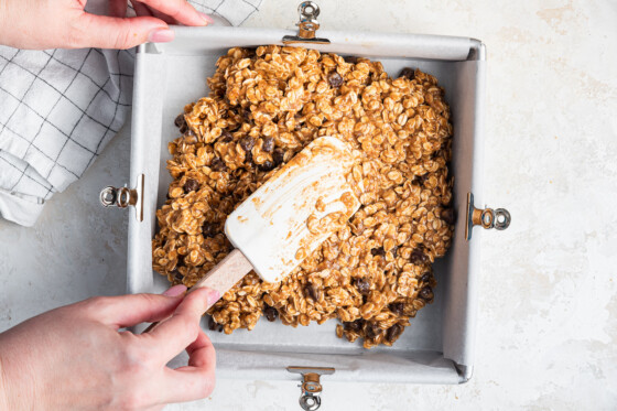 A woman's hand using a silicone spatula to form the oatmeal chocolate chip bars in a square baking pan.