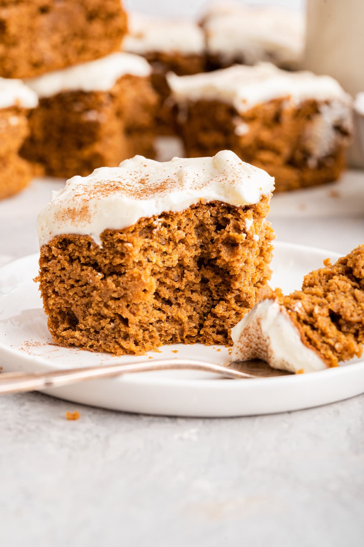 Pumpkin cake slice with a cream cheese frosting on a small white plate with a fork, taking a bite size piece from the slice.