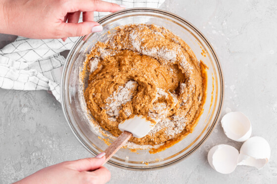 A woman's hand with a silicone spatula being used to mix the wet and dry ingredients for the healthy pumpkin cake in a large glass mixing bowl.