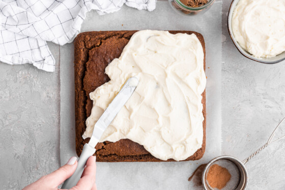 Healthy pumpkin cake on parchment paper being frosted using a metal spatula and a cream cheese frosting.