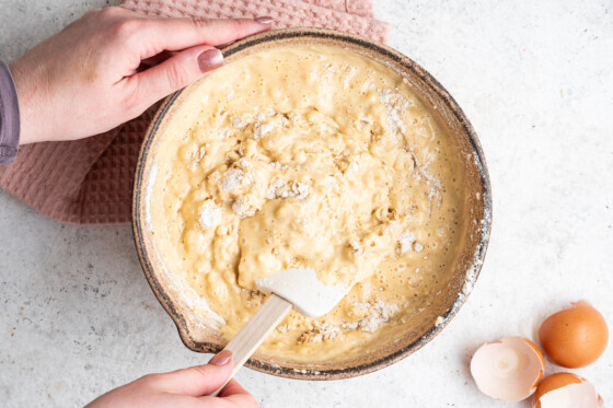 A woman's hand with a silicone spatula mixing the wet and dry ingredients for the vanilla cake in a large mixing bowl.