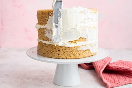 A spatula applying white frosting to three layers of vanilla cake that tis sitting on a white pedestal.