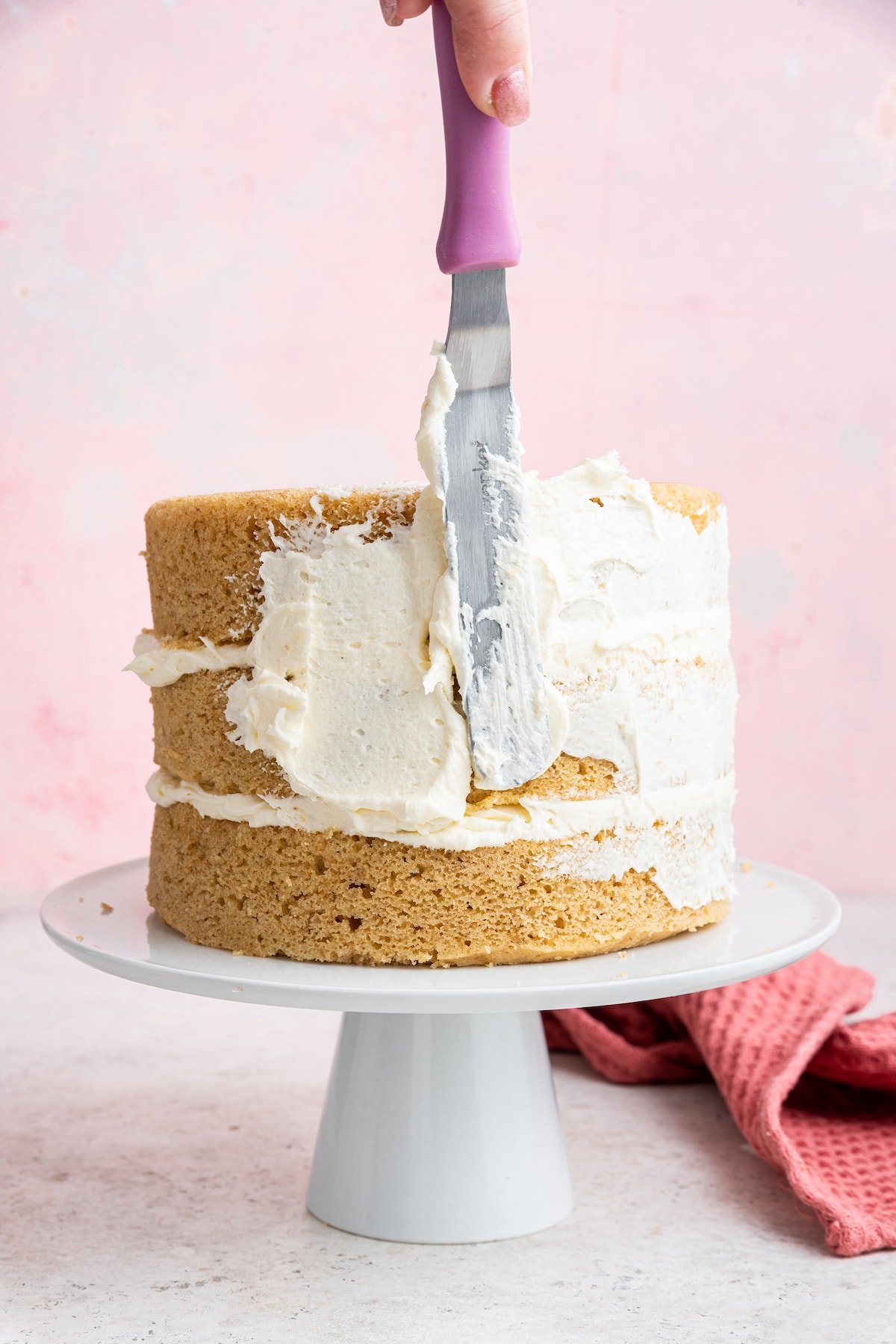 A woman's hand uses a spatula to apply white frosting to a vanilla cake.