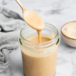 A hand holding a metal spoon with tahini over a glass jar full of tahini.