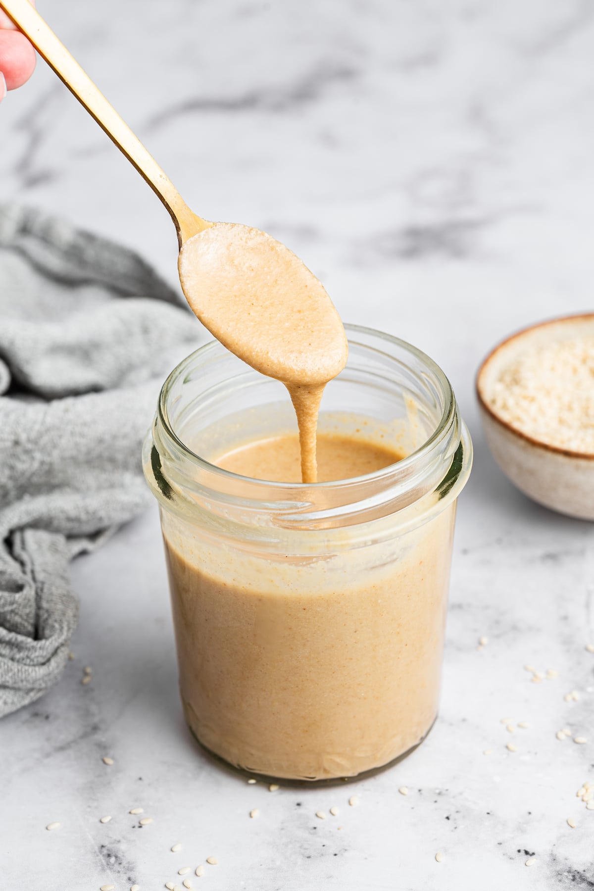 A hand holding a metal spoon with tahini over a glass jar full of tahini.