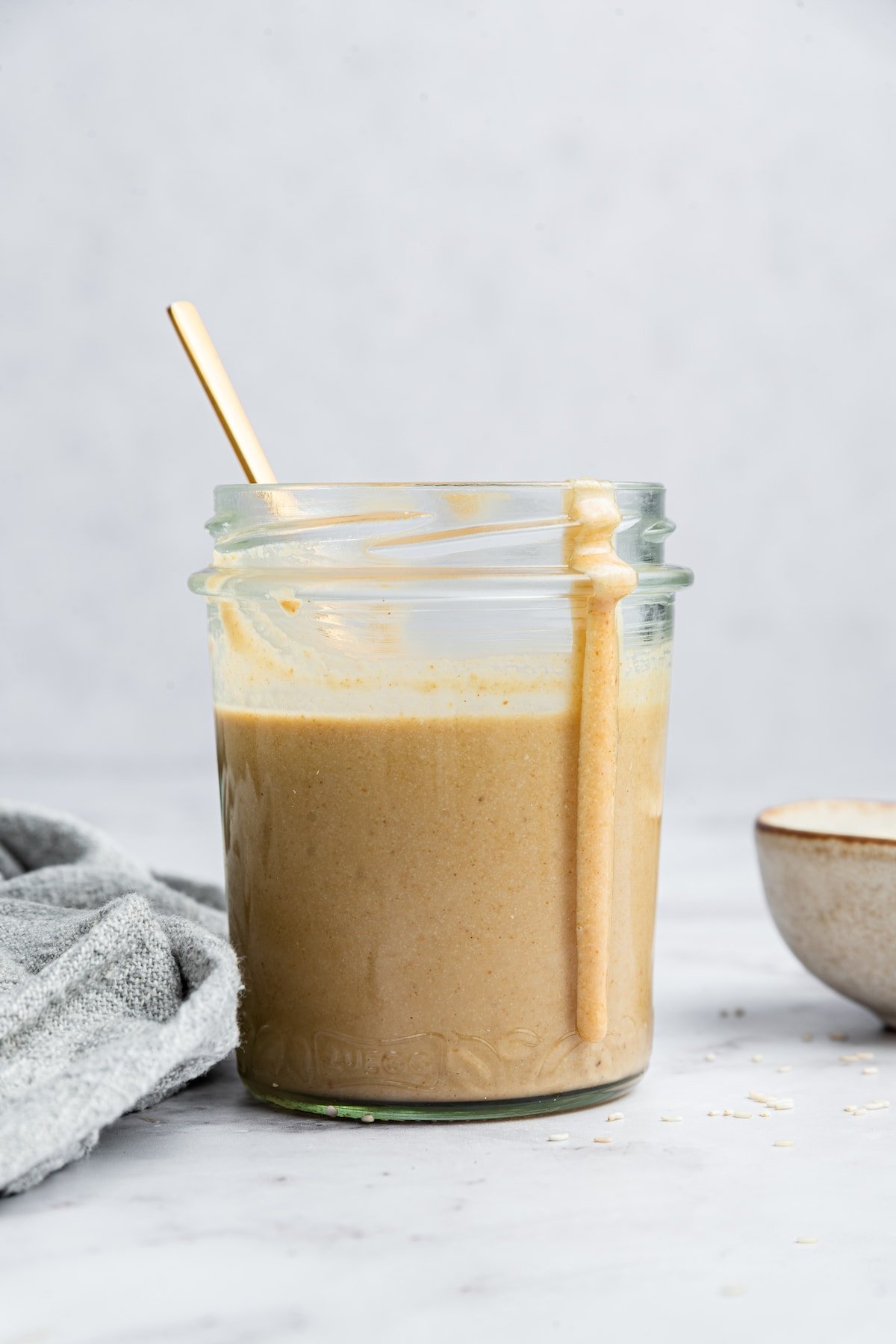 Tahini in a glass mason jar with a spoon inside and a stream of tahini on the outside of the jar.