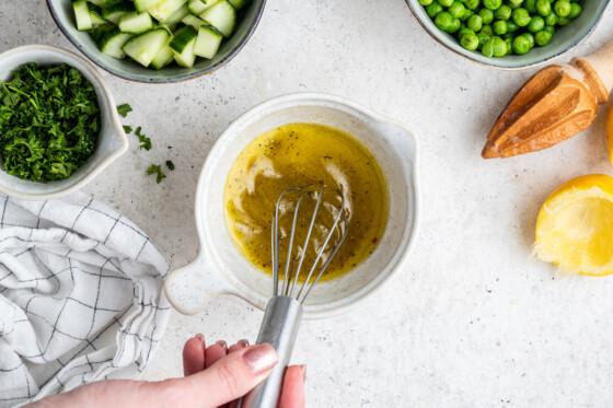 A woman's hand using a metal whisk in a small white bowl with lemon juice, olive oil, salt, and pepper.