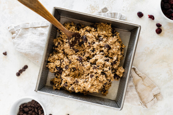 Dough for the oatmeal breakfast bars in a square baking pan with a large wooden spoon.