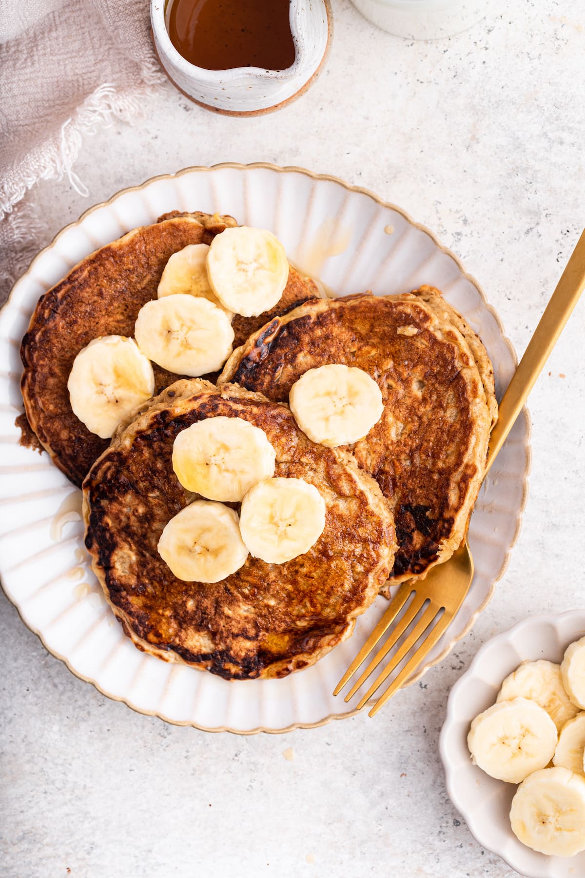 Three overnight oatmeal pancakes topped with banana slices and maple syrup on a plate with a fork.