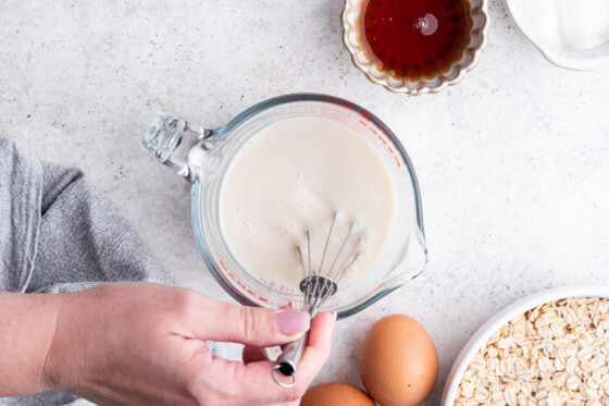 A woman's hand uses a metal whisk in a measuring cup of milk.