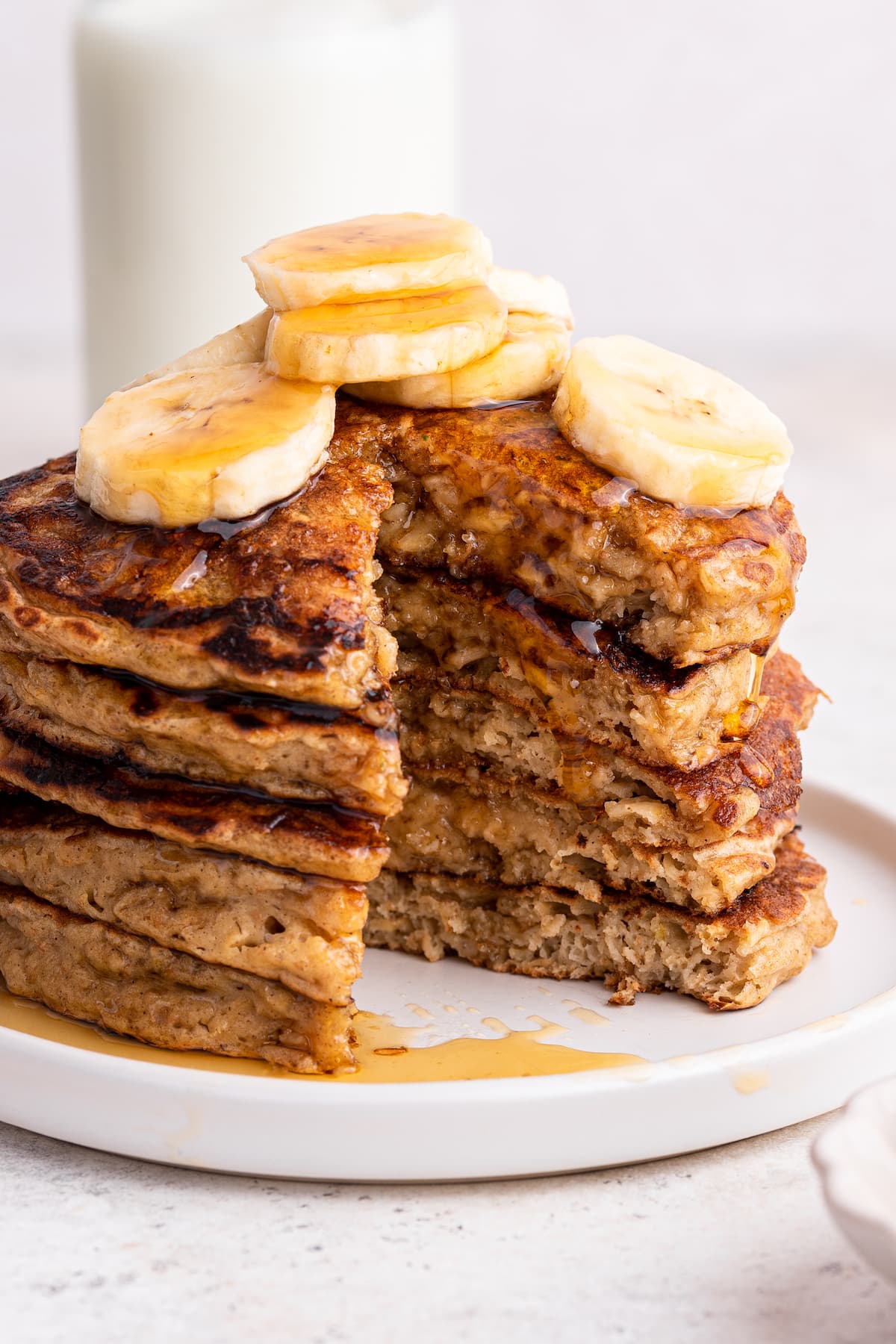 Five overnight oatmeal pancakes stacked on one another on a small plate with slices of banana and maple syrup. The stack of pancakes has a large triangular piece missing.
