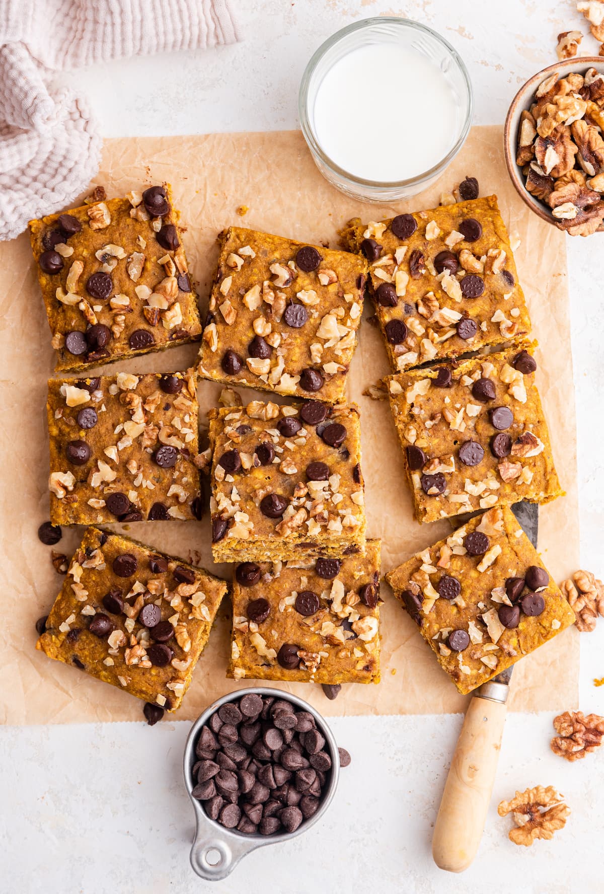 Pumpkin oatmeal bars on parchment paper near a glass of milk, a knife, a small bowl of walnuts, and a small bowl of chocolate chips.