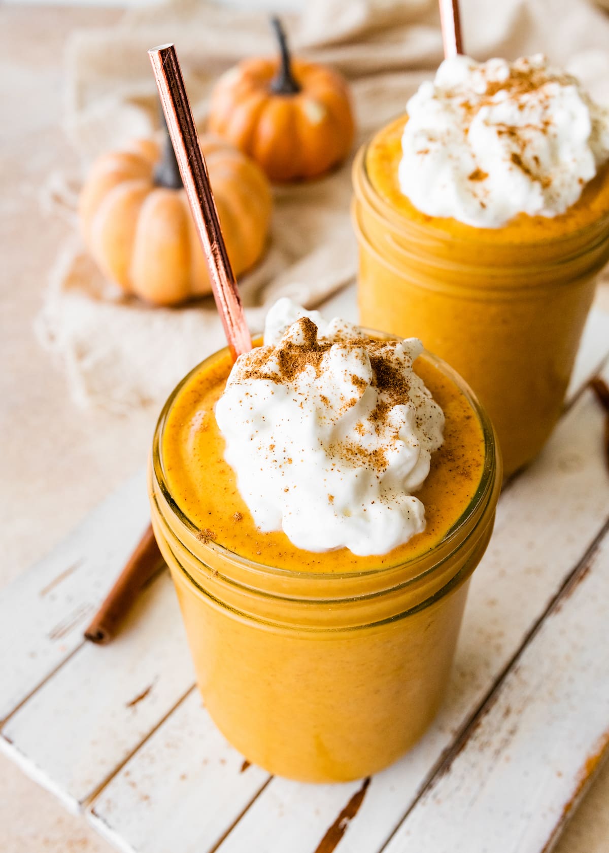 Two pumpkin protein shakes in glass mason jars with a metal straw, topped with whipped cream and ground cinnamon.