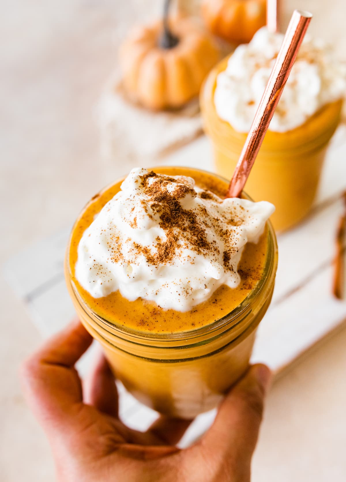 A hand holding a pumpkin protein shake in a glass jar topped with whipped cream and ground cinnamon.