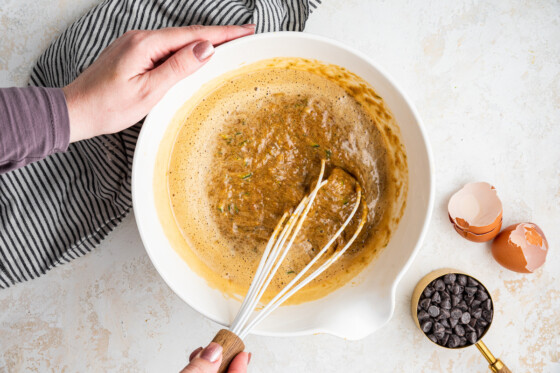 Woman's hands mixing the wet ingredients for the zucchini banana bread in a large white bowl using a whisk.