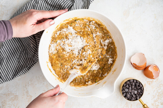 Woman's hands mixing the zucchini banana bread batter in a large white bowl using a spatula.