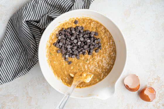 Zucchini banana bread batter in a large white bowl with a spatula and a cluster of chocolate chips.