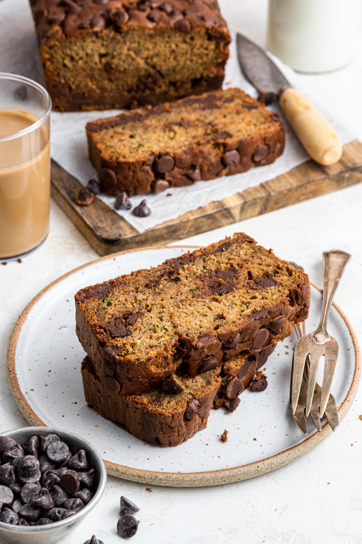 Two slices of zucchini banana bread stacked on a plate with the loaf of zucchini banana bread on a cutting board nearby.