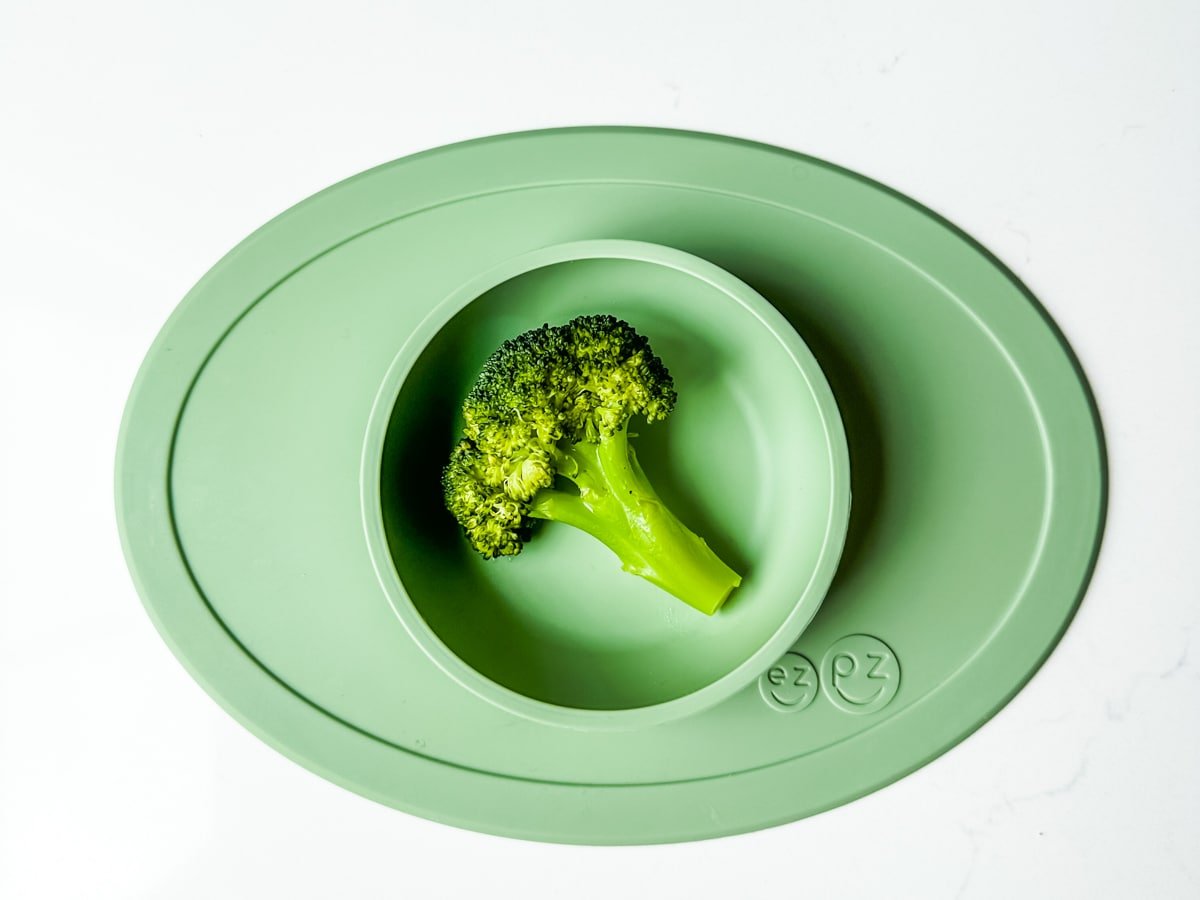 One piece of broccoli in a green ezpz baby bowl.