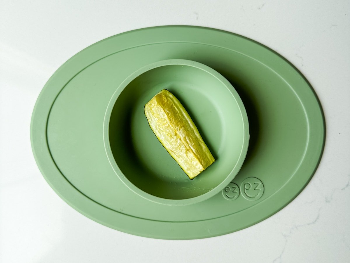 A piece of zucchini in a green ezpz baby bowl.