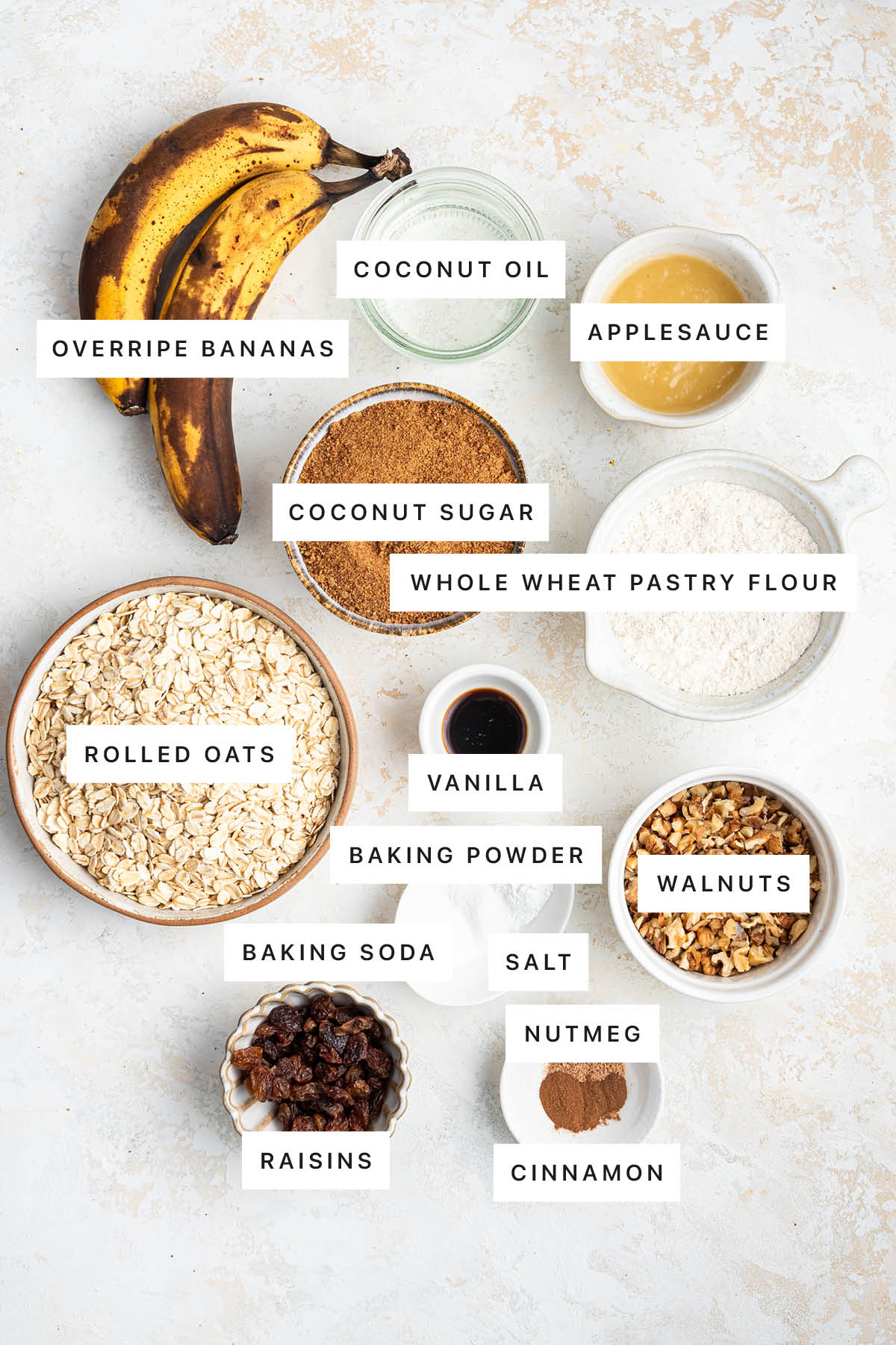 Ingredients measured out to make Healthy Banana Oat Bars: overripe bananas, coconut oil, applesauce, coconut sugar, whole wheat pastry flour, rolled oats, vanilla, baking powder, walnuts, baking soda, salt, nutmeg, raisins and cinnamon.