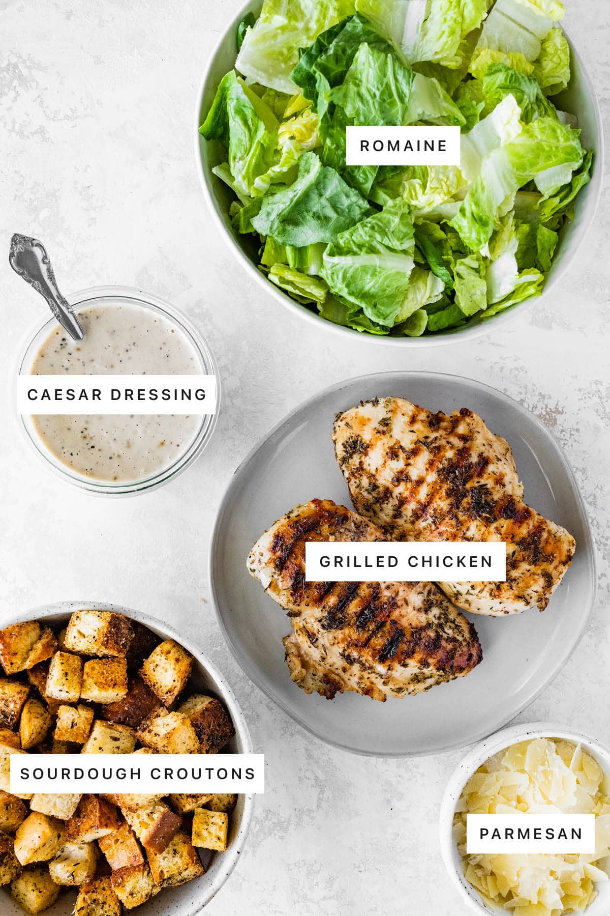 Ingredients measured out to make Grilled Chicken Caesar Salad: romaine, caesar dressing, grilled chicken, sourdough croutons and parmesan.