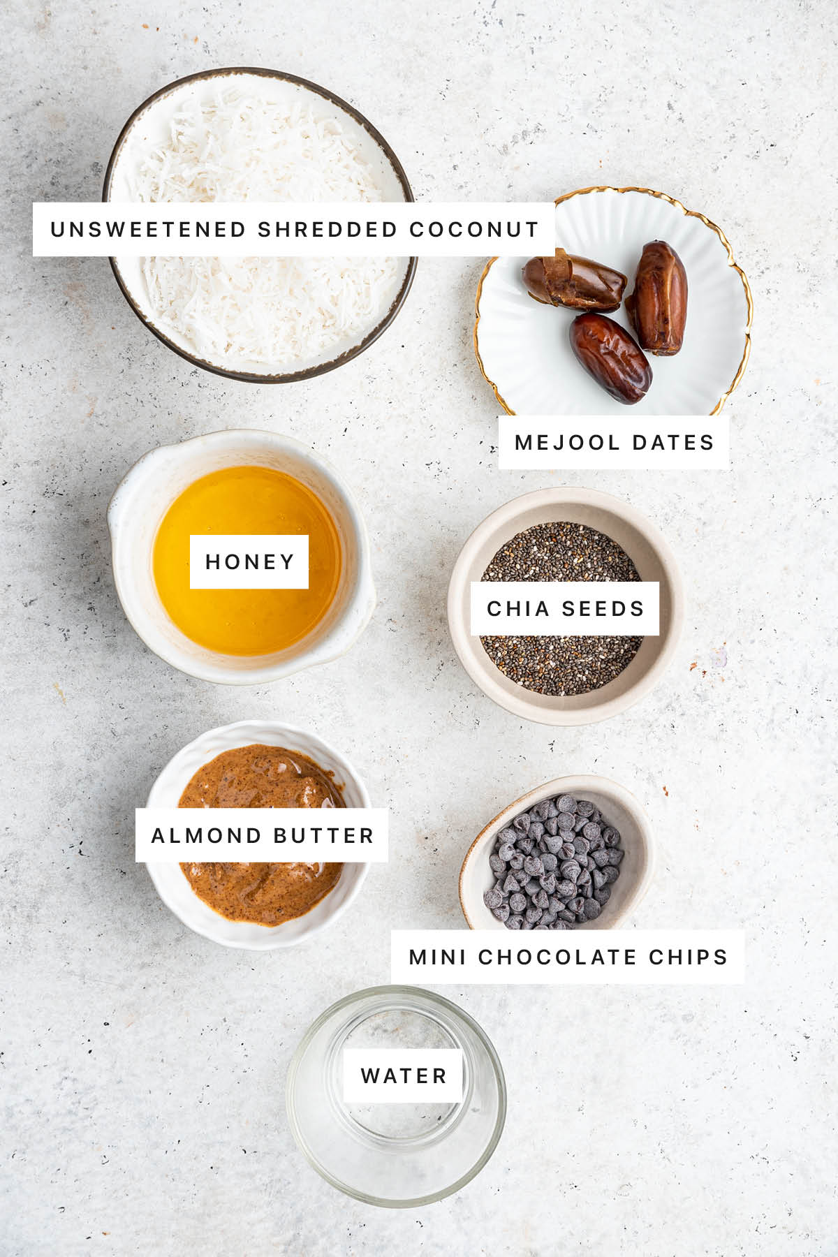 Ingredients measured out to make Toasted Coconut Chia Bars: unsweetened shredded coconut, medjool dates, honey, chia seeds, almond butter, mini chocolate chips and water.