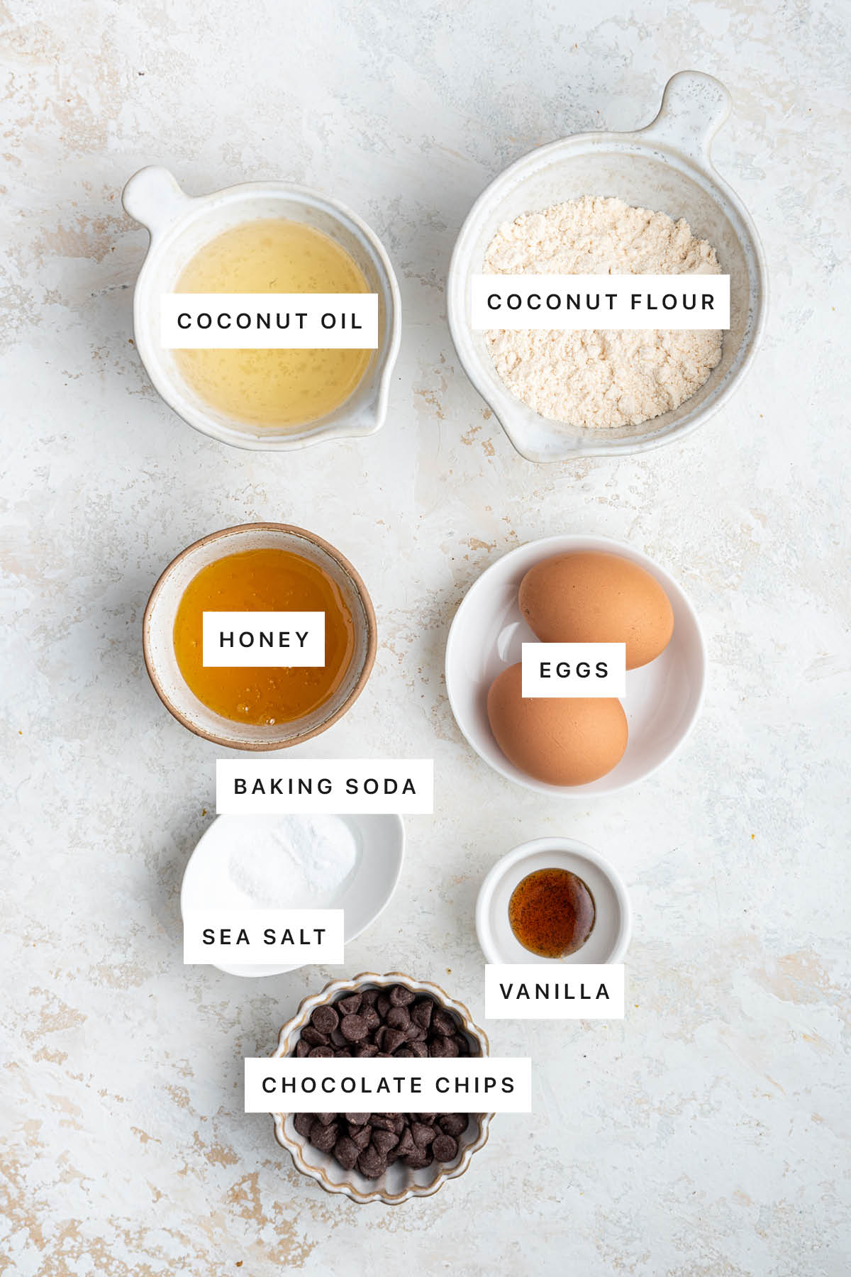Ingredients measured out to make Coconut Flour Cookies: coconut oil, coconut flour, honey, eggs, baking soda, sea salt, vanilla and chocolate chips.