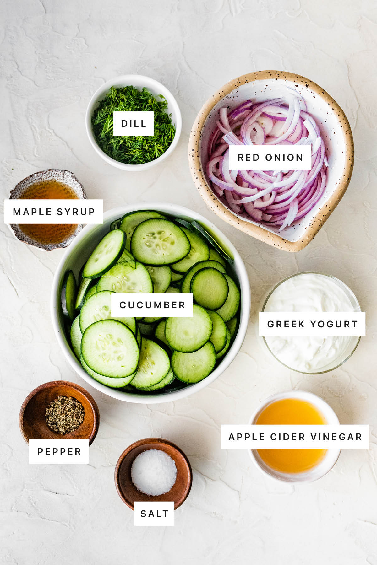 Ingredients measured out to make Creamy Cucumber Salad: dill, red onion, maple syrup, cucumber, Greek yogurt, pepper, salt and apple cider vinegar.