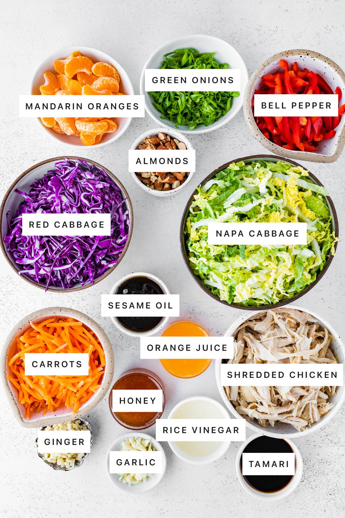 Ingredients measured out to make Mandarin Orange Chicken Salad: mandarin oranges, green onions, bell pepper, almonds, red cabbage, napa cabbage, sesame oil, orange juice, carrots, honey, shredded chicken, ginger, garlic, rice vinegar and tamari.